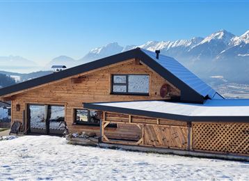 Ein modernes Holzhaus in einer schneebedeckten Landschaft. Im Hintergrund sind Berge und ein klarer blauer Himmel sichtbar.