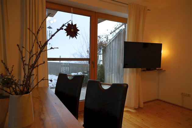 Ein gemütlicher Raum mit einem Holzstammtisch und schwarzen Stühlen. Durch das Fenster sieht man eine verschneite Landschaft und einen Fernseher an der Wand.