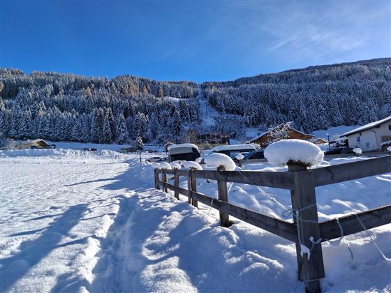 Een besneeuwd landschap met een houten hek en besneeuwde heuvels. Op de achtergrond zijn bossige bergen te zien onder een heldere blauwe lucht.