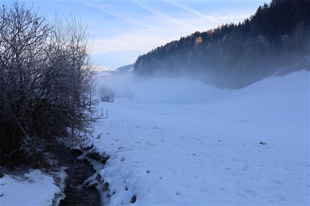 Eine verschneite Landschaft mit einem klaren Himmel und Nebel im Hintergrund. Ein kleiner Bach fließt durch die Szene.
