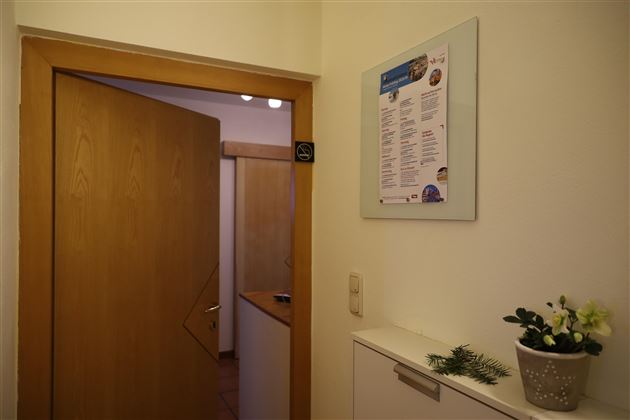 Ein Eingangsbereich mit einer Holz-Tür und einem Informationsplakat an der Wand. Auf der Anrichte stehen dekorative Pflanzen.
