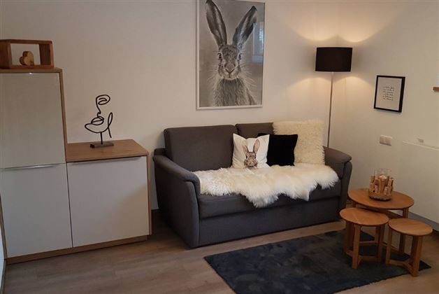A cozy living room with a gray sofa and a white blanket. On the wall hangs a picture of a rabbit, and there is a small table with decoration.