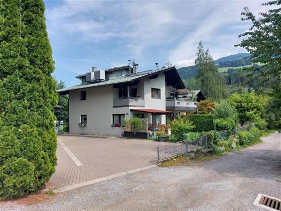 Ein modernes Wohnhaus umgeben von Bäumen und Grünflächen. Die Straße ist gepflastert und führt in eine ruhige Nachbarschaft.