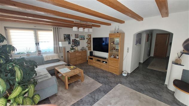Ein gemütliches Wohnzimmer mit Holzdecke und moderner Möbeln. Ein Sofa, ein Fernseher und Pflanzen schaffen eine einladende Atmosphäre.